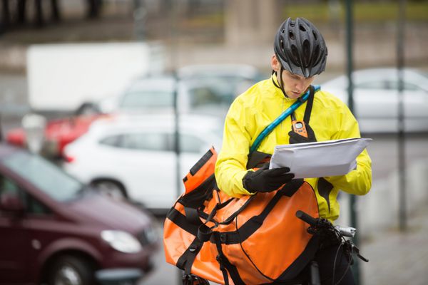 دوچرخه سوار مرد جوان با وسایل محافظ با بسته و کیف پیک در خیابان