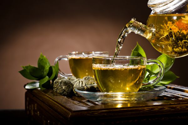 پو افقی از قوری شیشه ای جریان چای سبز در فنجان در زمینه قهوه ای مراسم چای