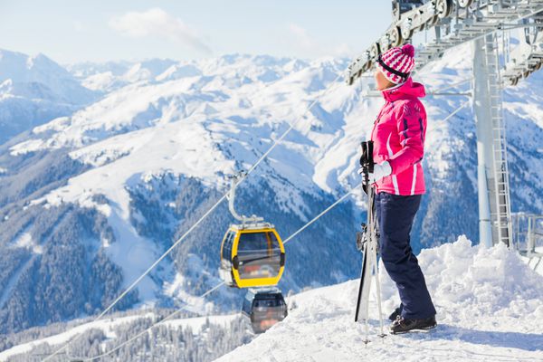 زن خندان شاد با عینک اسکی در مقابل یک تله اسکی و پس زمینه کوه های زمستانی فوق العاده در پس زمینه کوه های زمستانی شگفت انگیز و زلرتال اتریش