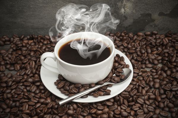 فنجان قهوه صبحانه و نعلبکی احاطه شده با دانه های قهوه در پس زمینه روستایی