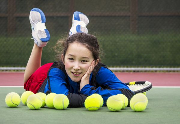 پرتره افقی دختر نوجوان تنیس باز خندان که در زمین بیرونی پشت ردیفی از توپ های تنیس دراز کشیده و سر در دست و پاهایش در هوا قرار گرفته است