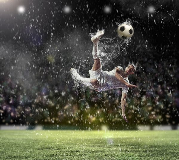 بازیکن فوتبال با پیراهن سفید در حال ضربه زدن به توپ در استادیوم زیر باران