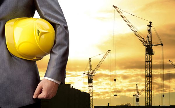 مهندس با در دست داشتن کلاه زرد برای امنیت کارگران در پس زمینه ساختمان های آپارتمانی جدید و جرثقیل های ساختمانی در پس زمینه غروب آفتاب شب آسمان ابری جرثقیل بار را بلند می کند