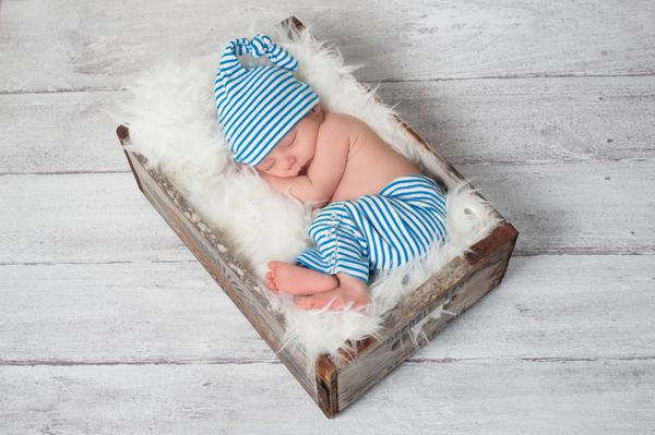 نوزاد تازه متولد شده ای که لباس خواب راه راه آبی و سفید پوشیده و در یک جعبه چوبی قدیمی و قدیمی می خوابد