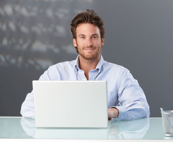 تاجر کاس با کامپیوتر لپ تاپ در دفتر نشسته و به دوربین لبخند می زند
