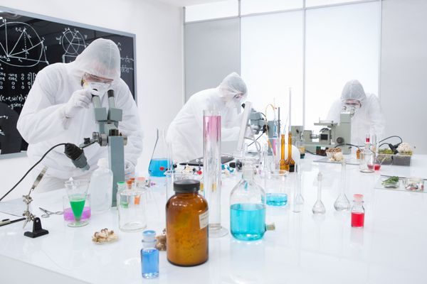 نمای کلی سه نفر در حال مشاهده و تجزیه و تحلیل واکنش های شیمیایی در آزمایشگاه با استفاده از مواد رنگارنگ و ابزار آزمایشگاهی روی میز کار آزمایشگاه با تخته سیاه در پس زمینه