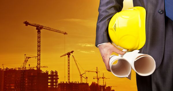 کلاه زرد مهندسی برای امنیت کارگران در پس‌زمینه ساختمان‌های آپارتمانی مرتفع و جرثقیل‌های ساختمانی و غروب آفتاب عصر