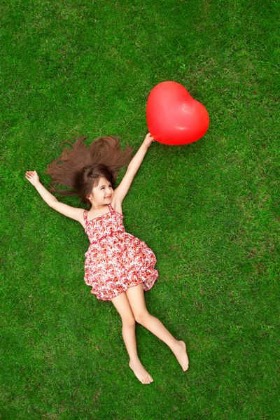 دختر زیبا با لباس رنگی که روی چمن دراز کشیده و یک توپ قرمز به شکل قلب در دست گرفته است