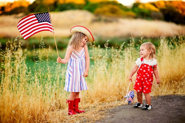 گروهی متشکل از دو دختر بچه کوچولوی شاد و شایان ستایش که در بیرون لبخند می زنند و پرچم آمریکا را تکان می دهند لباس او با نوار و ستاره کلاه گاوچران کودک خندان چهارم ژوئیه - روز استقلال را جشن می گیرد