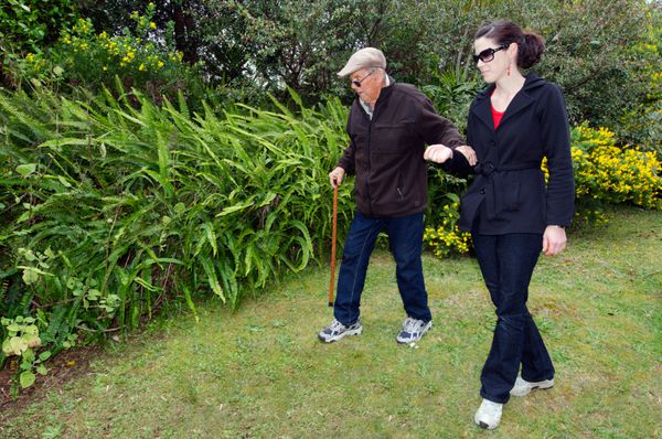 زن جوان در حال کمک و حمایت از پدربزرگش برای راه رفتن در فضای باز در باغ با چوب دستی او