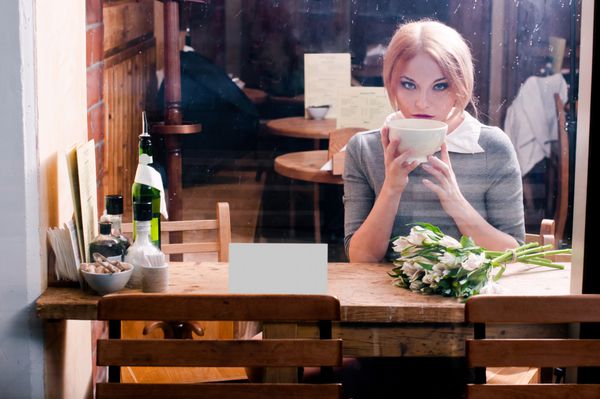 زن با فنجان قهوه با کت و شلوار خاکستری پشت پنجره کافه