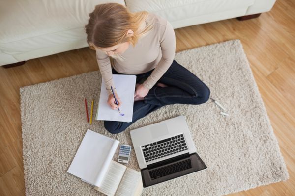 زن جوان در حال انجام تکالیف و نشستن روی زمین با استفاده از لپ تاپ در خانه