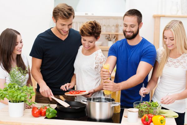 گروهی از دوستان جوان متنوع در حال آماده کردن شام با هم در آشپزخانه ایستاده پشت پیشخوان و پختن اسپاگتی و سالاد