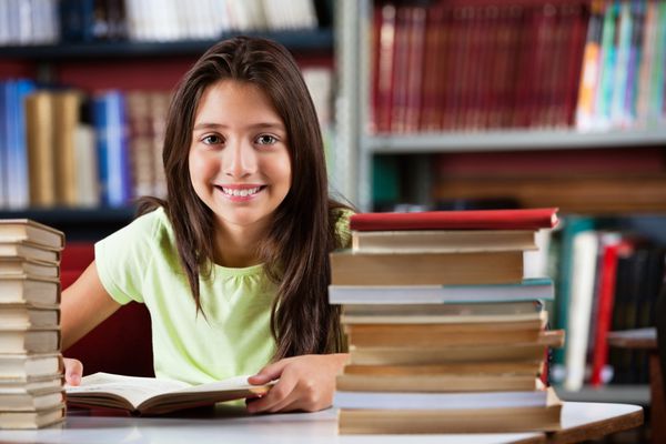 پرتره دختر مدرسه ای ناز در حالی که با پشته کتاب پشت میز کتابخانه نشسته است لبخند می زند