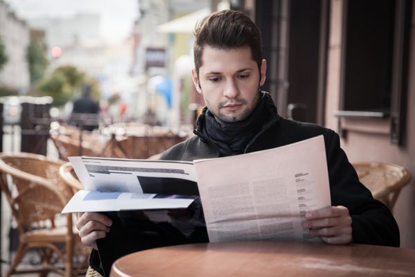 مرد جوان خوش تیپ در حال خواندن مجله در کافه ای در خیابان