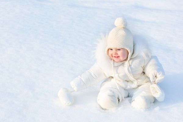 دختر بچه ی زیبا روی برف سفید نشسته و ژاکت گرم و کلاه بافتنی به تن دارد