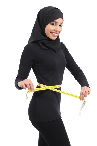 زن زیبای تناسب اندام عرب عربستانی که دور کمر خود را با متر اندازه می گیرد و به دوربین جدا شده در پس زمینه سفید نگاه می کند