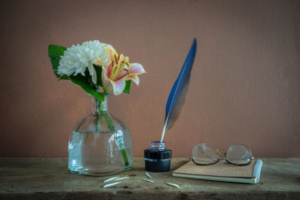 طبیعت بی جان با گل محمدی سفید در بطری قلم پر در بطری جوهر و شیشه قدیمی روی کتاب قهوه ای
