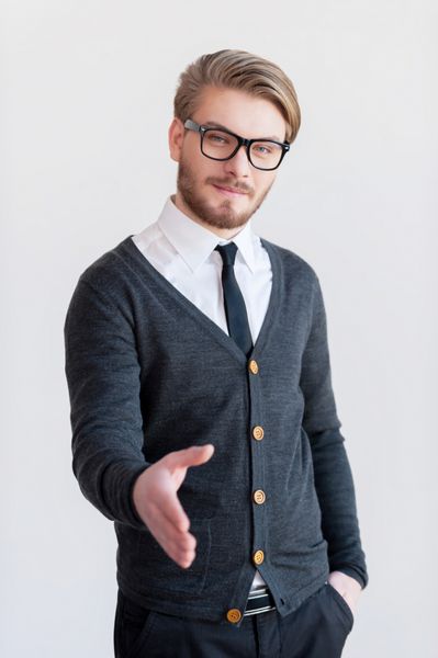 به عرشه خوش آمدید مرد جوان خوش تیپ با عینک در حالی که در مقابل پس زمینه خاکستری ایستاده است برای تکان دادن دستش را دراز می کند