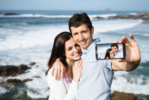 زوج جوان در سفر ماه عسل در سواحل آستوریاس اسپانیا در حال گرفتن عکس پرتره با دوربین گوشی هوشمند