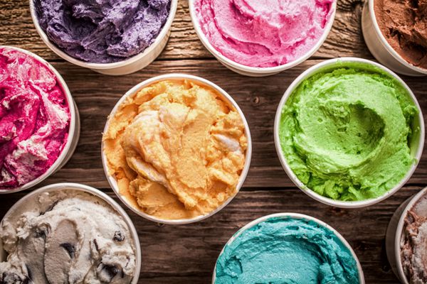 مجموعه ای از بستنی های رنگارنگ با طعم های مختلف در وان های پلاستیکی که روی میز چوبی قدیمی در یک بستنی فروشی برای تنقلات یخ زده خوشمزه در یک روز تابستانی نمایش داده شده است