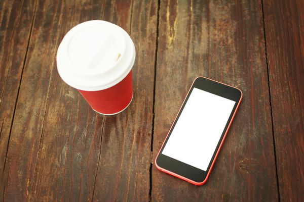 نمای نزدیک از تلفن هوشمند با صفحه سفید جدا شده و فنجان قهوه آماده روی میز چوبی مفهوم ماکت