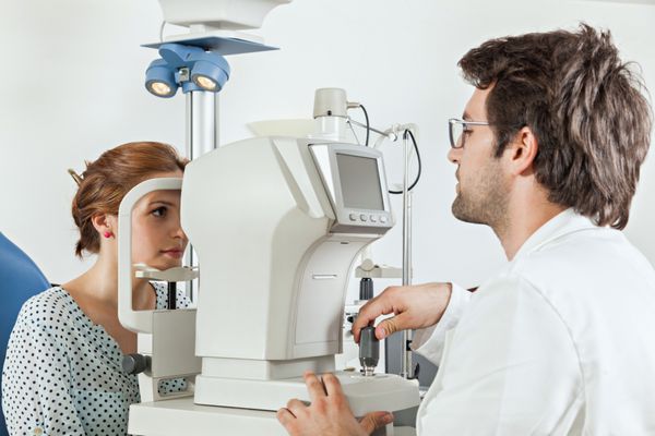 چشم پزشک در اتاق معاینه با زن جوانی که روی صندلی نشسته و به دستگاه تست چشم نگاه می کند