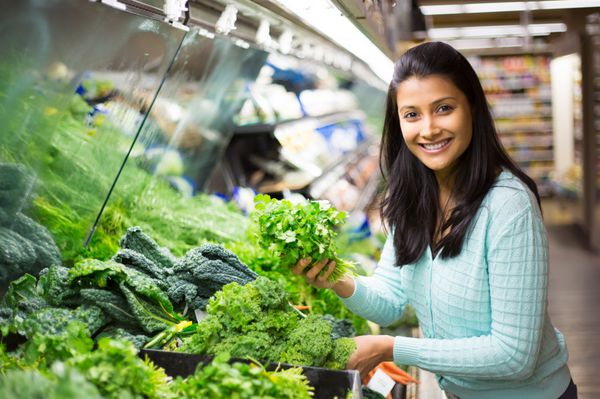 پرتره نزدیک زن جوان زیبا و زیبا با ژاکت در حال برداشتن انتخاب سبزیجات برگ سبز در فروشگاه مواد غذایی