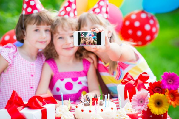 کودکان شادی که با گوشی هوشمند سلفی می گیرند جشن تولد در فضای باز