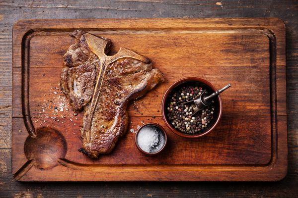 استیک تی بون کبابی با نمک و فلفل روی تخته برش گوشت در زمینه چوبی تیره