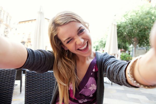 پرتره نزدیک از یک زن جوان جذاب که یک دوربین دیجیتال گوشی هوشمند را با دستان خود در دست گرفته و یک سلفی سلفی از خود در حال گرفتن چشمک به دوربین می گیرد سفر و فناوری در فضای باز