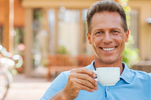 زمان استراحت مرد بالغ شاد در حال نوشیدن قهوه و لبخند زدن در حالی که بیرون از خانه نشسته و خانه در پس‌زمینه است