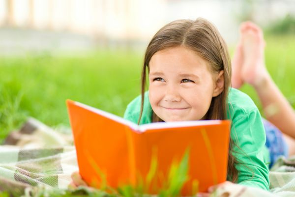 دختر کوچولوی ناز در حالی که روی چمن سبز دراز کشیده در حال خواندن کتاب است