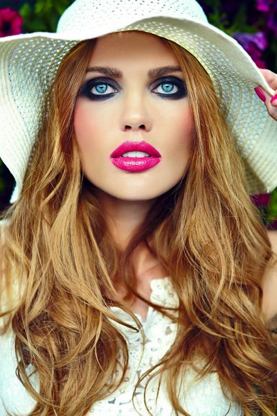 ظاهر مد بالا پرتره نزدیک از مدل زن جوان زیبا و شیک با آرایش روشن و لب های صورتی با پوست تمیز کامل در کلاه نزدیک گل های تابستانی