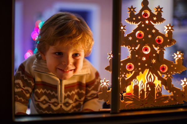پسر ناز خندان که هنگام کریسمس کنار پنجره ایستاده است با چراغ های رنگارنگ از درخت کریسمس در پس زمینه فوکوس انتخابی