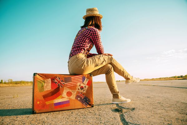 زن مسافر روی چمدان رترو می نشیند و به جاده نگاه می کند چمدان با تمبر پرچم هایی که نشان دهنده هر کشور سفر شده است