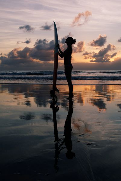 دختر جوان موج سوار با تخته موج سواری در ساحل هنگام غروب آفتاب