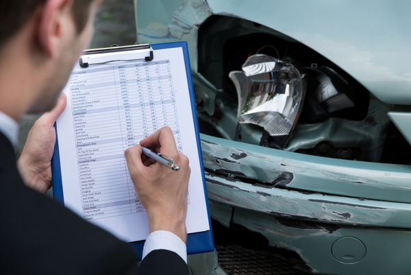 نمای جانبی نوشتن روی کلیپ بورد در حالی که نماینده بیمه در حال معاینه خودرو پس از تصادف است
