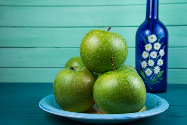 مزرعه سیب سبز تازه روی میز چوبی و بطری با گل در پس زمینه پاستل