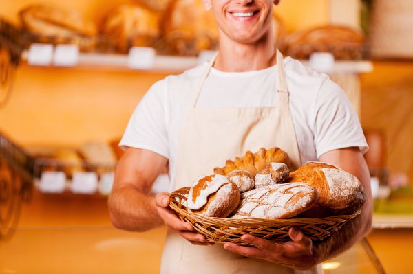 تازه ترین نان برای شما تصویر برش خورده مرد جوان در پیش بند که سبد با محصولات پخته شده در دست دارد و در حالی که در مغازه نانوایی ایستاده لبخند می زند