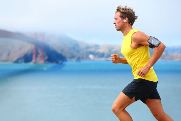 مرد ورزشکار - دونده مرد در سانفرانسیسکو در حال گوش دادن به موسیقی از طریق تلفن هوشمند مرد جوان خوش اندام در حال دویدن در کنار خلیج سان فرانسیسکو و پل گلدن گیت آموزش دویدن با بازوبند تلفن هوشمند