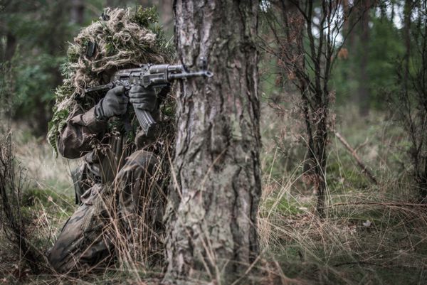 سرباز پوشیده در لباس گیلی با تفنگ تهاجمی پنهان شده در پشت درخت - نمای جلو