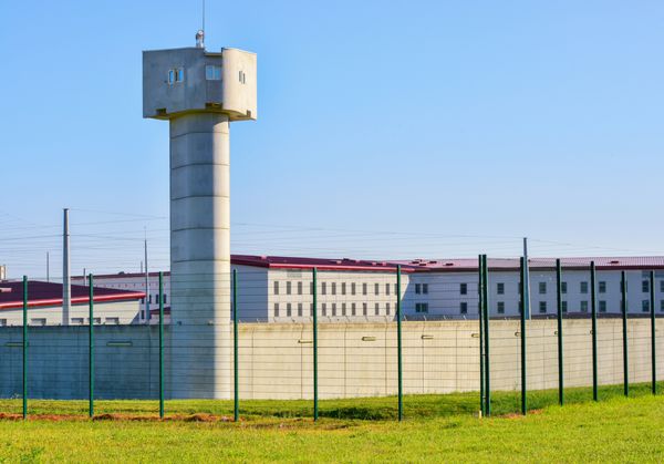 محوطه زندان بزرگ با برج مراقبت بلند که با حصار احاطه شده و آسمان آبی در پس زمینه