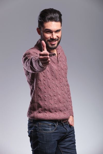 مرد جوان مد و خندان در حالی که یک دستش را در جیب خود گرفته است حرکت شست را به سمت بالا نشان می دهد در زمینه خاکستری