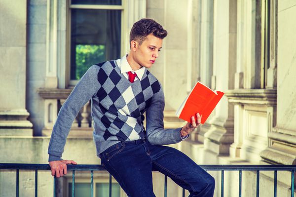 مردی که بیرون می خواند با یک ژاکت طرح دار مشکی سفید خاکستری شلوار جین یک دانشجوی جوان خوش تیپ روی نرده ای در ساختمان اداری نشسته و به طور متمرکز مشغول خواندن یک کتاب قرمز است