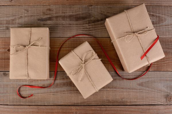 زاویه بالا از سه هدیه کریسمس پیچیده شده ساده کاغذ قهوه‌ای و بسته‌های بسته‌بندی شده با روبان قرمزی که از وسط آن روی موج‌سواری چوبی روستایی می‌گذرد
