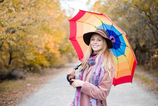 پرتره بانوی جوان زیبا با چتر رنگارنگ رنگین کمانی در حال سرگرمی و لبخند زدن شاد و نگاه کردن به دوربین در جاده پاییزی در برابر پس زمینه کپی آسمان روشن