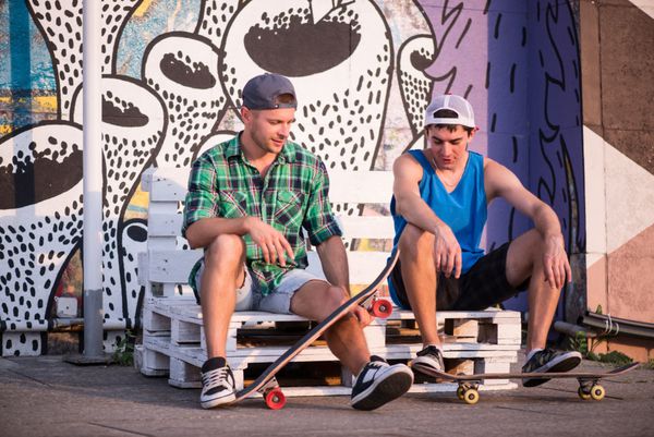 پرتره تمام قد از دو مرد جوان که روی نیمکت سفید در خیابان نشسته اند و اسکیت بردهای خود را به یکدیگر نشان می دهند و در مورد فعالیت ورزشی مورد علاقه خود صحبت می کنند