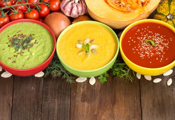 سه سوپ تازه در کاسه های رنگارنگ و سبزیجات روی میز چوبی