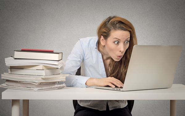 زن با استفاده از رایانه لپ‌تاپ خود پشت میز با انبوهی از کتاب‌ها در پس‌زمینه اداری دیوار خاکستری نشسته است مفهوم شیدایی اعتیاد به اینترنت با فناوری جدید دختری بامزه و شیفته کامپیوتر احساسات انسانی